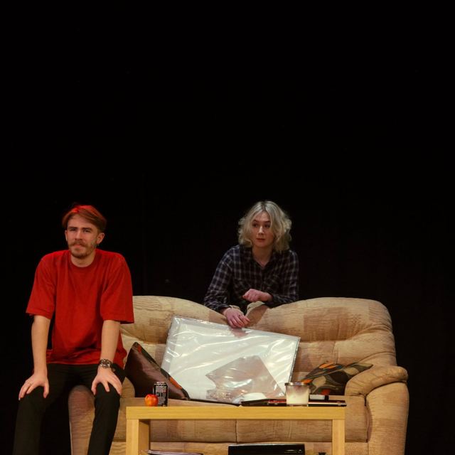A young man in a red T-shirt on a sofa beside a friend