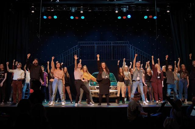 The company dancing around a car on stage