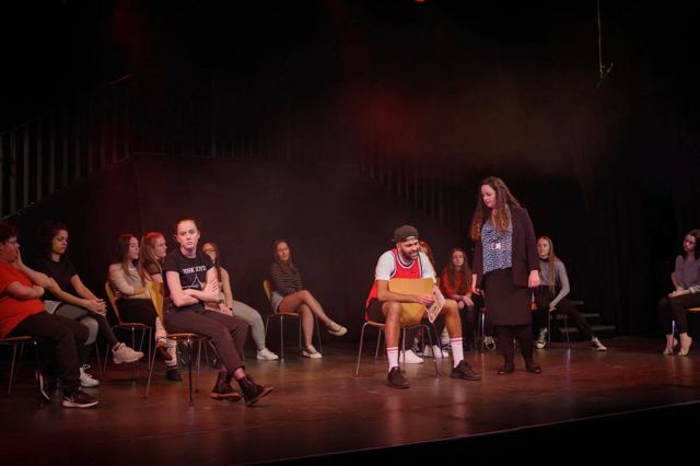 The cast sitting in a circle of chairs on stage
