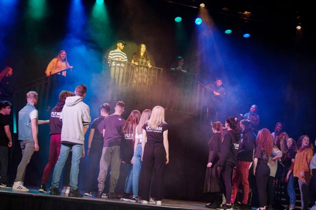 The cast under coloured stage lights, with performers on the platform above