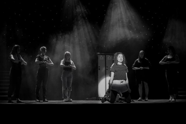Black-and-white photo of a performer kneeling in a spotlight, the cast behind her