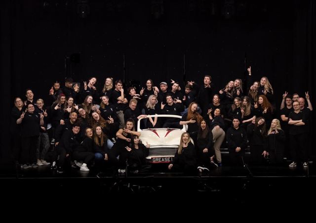 The whole company posing on stage around a white car