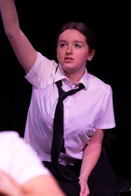A performer in a school shirt and tie with her arm raised