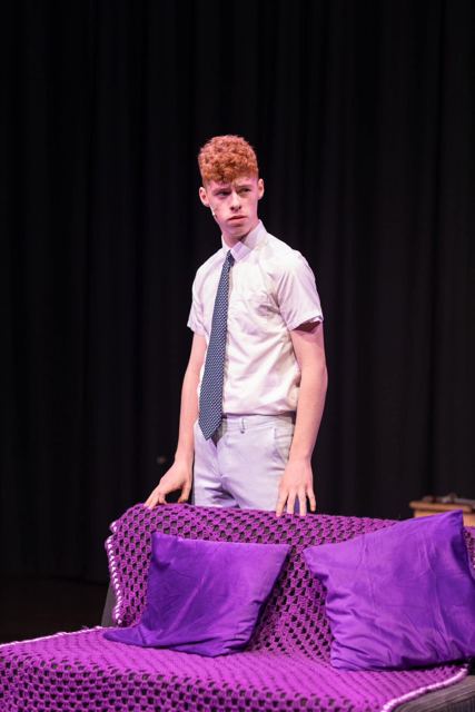 A young man in a school shirt standing behind a purple sofa