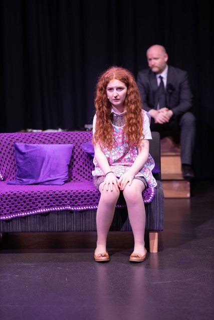 A young woman on a purple sofa, a man in black watching