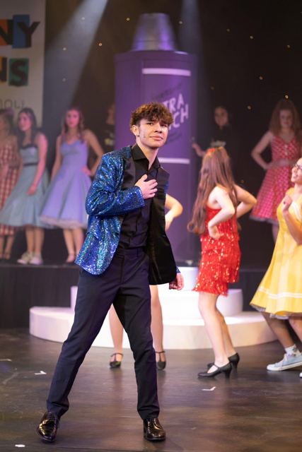 A performer in a sparkly blue jacket dancing in front of the chorus