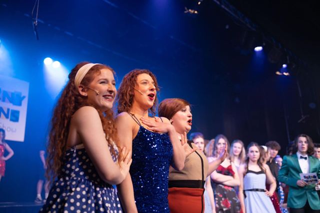 Performers in 1960s dresses singing together under blue light