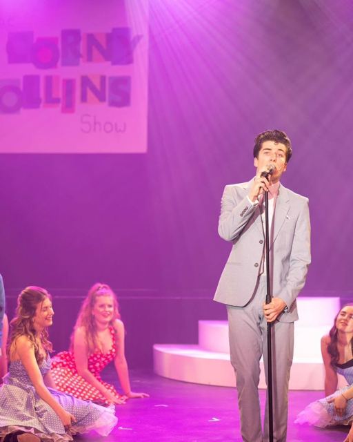 A presenter in a grey suit singing into a microphone, dancers at his feet