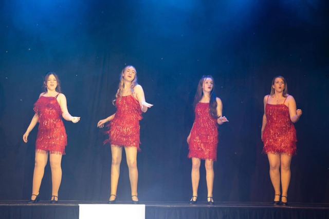 Four performers in red sequinned dresses singing on a raised platform