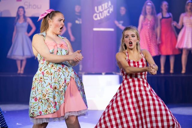 Two performers in floral and gingham dresses looking surprised