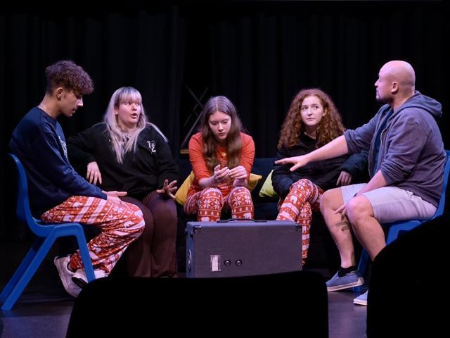 Performers sitting in a circle of chairs around a speaker