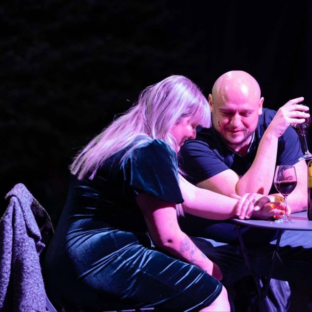 A woman and a man leaning close together at a small table with wine