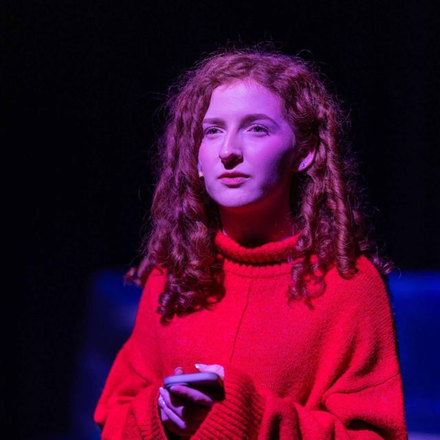 A young woman with curly hair in a red jumper, holding a phone