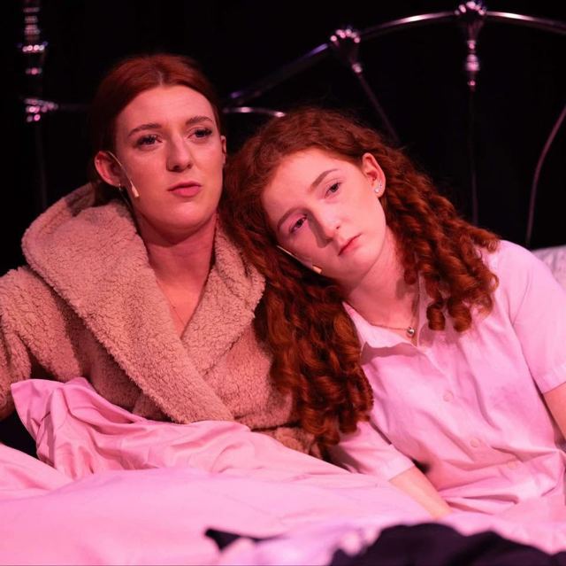 Two young women sitting up in bed, one resting her head on the other's shoulder