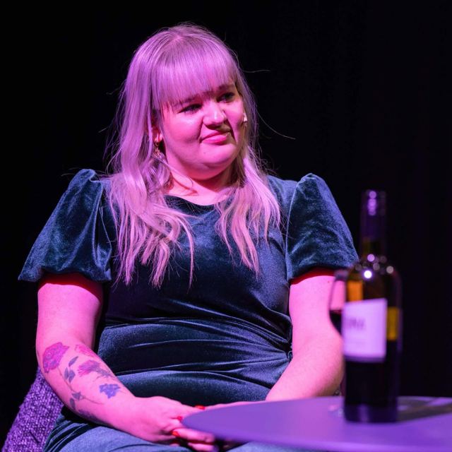 A woman with long blonde hair in a green velvet dress beside a bottle of wine