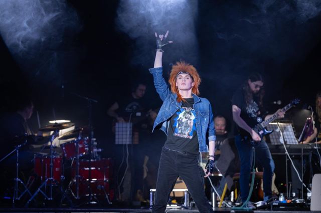 A performer in a denim jacket punches the air in front of the live band