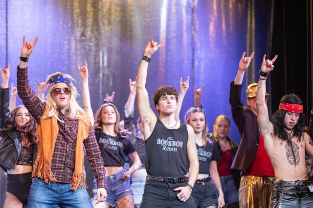 Cast members in denim, fringes and band T-shirts making rock hand signs