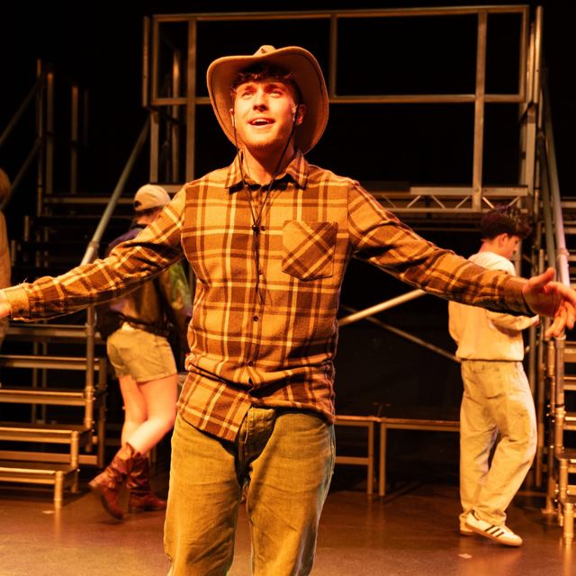 A performer in a plaid shirt and cowboy hat, arms wide