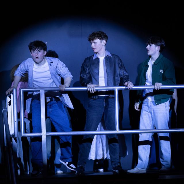 Three young men leaning on a scaffold rail in a spotlight