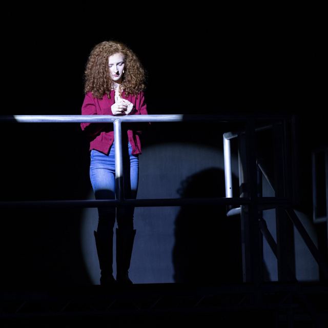 A performer in a red jacket at a rail in a spotlight