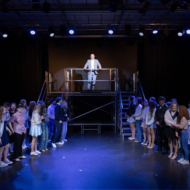 A wide view of the stage with the company and a figure on the platform above