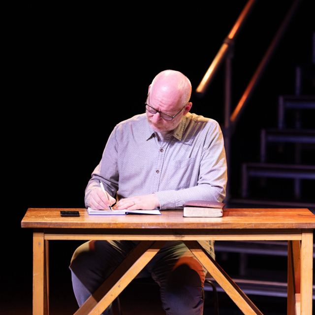 A man in a grey shirt writing at a wooden desk